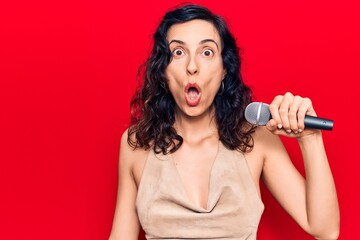 Young beautiful hispanic woman singing song using microphone scared and amazed with open mouth for...