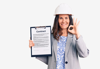 Young beautiful brunette woman wearing architect hardhat holding contract doing ok sign with fingers, smiling friendly gesturing excellent symbol