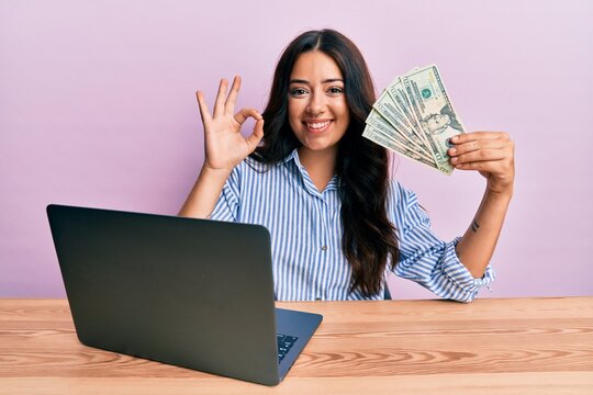 Beautiful brunette young woman working at the office holding dollars doing ok sign with fingers, smiling friendly gesturing excellent symbol
