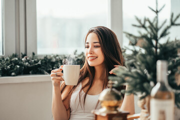 Brunette young asian woman in casual white shirt and jeans sitting in flat smiling happily with a cup of coffee in the morning. Lifestyle portrait indoors. Copy space