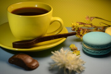 Obraz na płótnie Canvas A cup of morning coffee in a yellow mug with a chocolate spoon, blue macaroon and machine shaped chocolate near the flowers