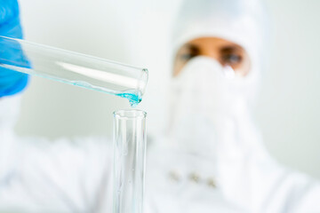 Young attractive concentrated female scientist in protective eyeglasses, mask and gloves dropping a blue liquid substance into the test tube with a pipette in the scientific chemical laboratory   