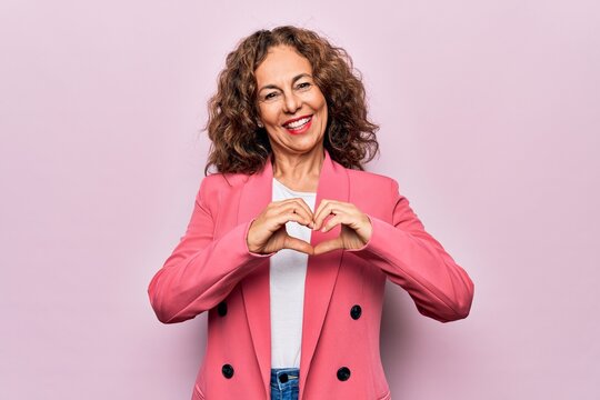 Middle age beautiful businesswoman wearing jacket standing over isolated pink background smiling in love doing heart symbol shape with hands. Romantic concept. - Powered by Adobe