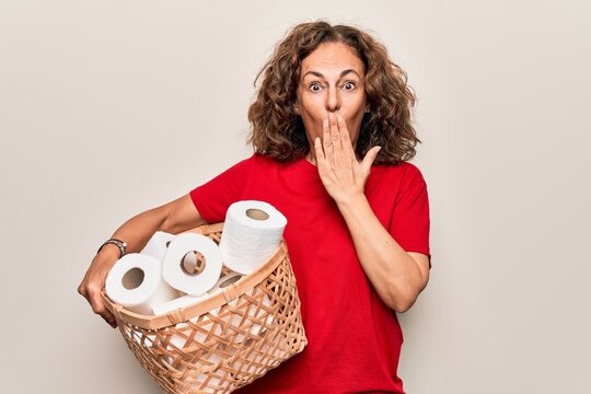 Middle age woman holding wicker basket with lots of toilet paper rolls over white background covering mouth with hand, shocked and afraid for mistake. Surprised expression