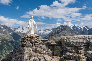 White statue of Virgin Mary, Mother of God, placed on top of the mountain. In the background there are snowy peaks of high mountains, blue sky, white clouds. - 396197321