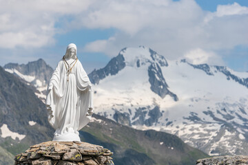 White statue of Virgin Mary, Mother of God, placed on top of the mountain. In the background there are snowy peaks of high mountains, blue sky, white clouds. - 396197365
