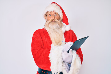 Old senior man with grey hair and long beard wearing santa claus costume writting list puffing cheeks with funny face. mouth inflated with air, catching air.