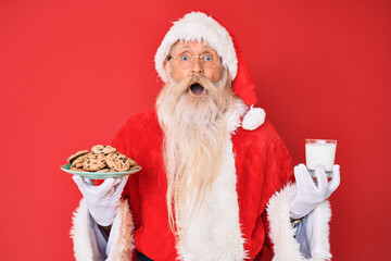 Old senior man with grey hair and long beard wearing santa claus costume holding cookies and milk afraid and shocked with surprise and amazed expression, fear and excited face.