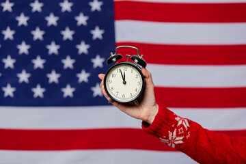 Female hand in Christmas sweater holds alarm clock on background with USA flag