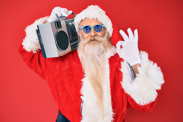 Old senior man wearing santa claus costume and boombox doing ok sign with fingers, smiling friendly gesturing excellent symbol
