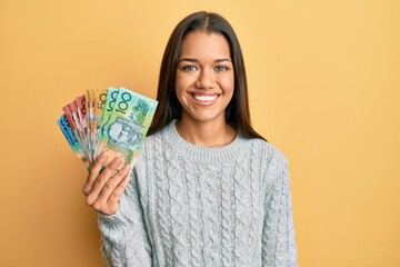 Beautiful hispanic woman holding australian dollars looking positive and happy standing and smiling with a confident smile showing teeth