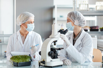 Portrait of two young female scientists looking in microscope while studying plant samples in...