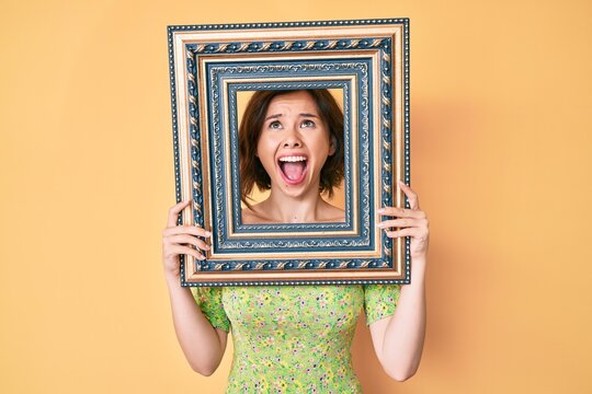 Young beautiful woman holding empty frame angry and mad screaming frustrated and furious, shouting with anger looking up.
