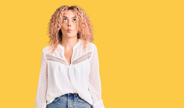 Young blonde woman with curly hair wearing elegant summer shirt making fish face with lips, crazy and comical gesture. funny expression.
