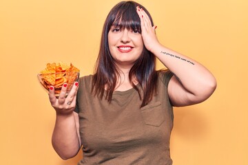 Young plus size woman holding nachos potato chips stressed and frustrated with hand on head, surprised and angry face