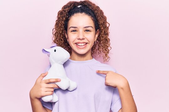 Beautiful kid girl with curly hair holding animal doll toy pointing finger to one self smiling happy and proud