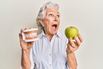 Senior grey-haired woman holding green apple and denture teeth angry and mad screaming frustrated...
