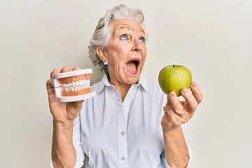 Fotobehang Senior grey-haired woman holding green apple and denture teeth angry and mad screaming frustrated and furious, shouting with anger looking up. © Krakenimages.com