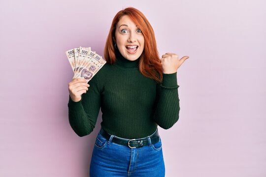 Beautiful redhead woman holding 500 mexican pesos banknotes pointing thumb up to the side smiling happy with open mouth