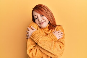 Beautiful redhead woman wearing casual winter sweater over yellow background hugging oneself happy and positive, smiling confident. self love and self care