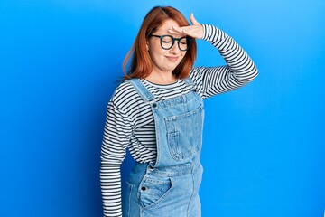 Beautiful redhead woman wearing casual clothes and glasses surprised with hand on head for mistake,...