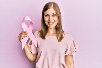 Beautiful caucasian woman holding pink cancer ribbon looking positive and happy standing and...