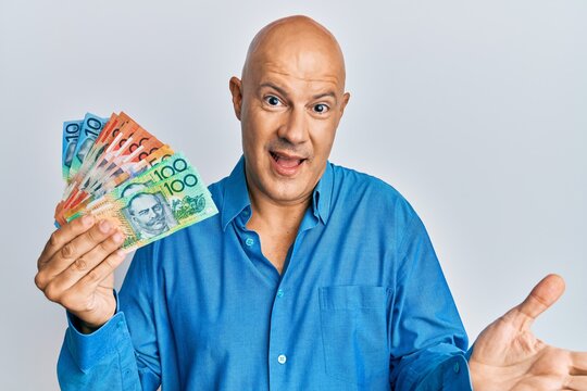Middle age bald man holding australian dollars celebrating achievement with happy smile and winner expression with raised hand