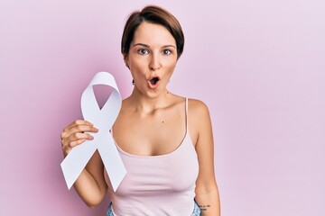 Young brunette woman with short hair holding cancer awareness white ribbon scared and amazed with...