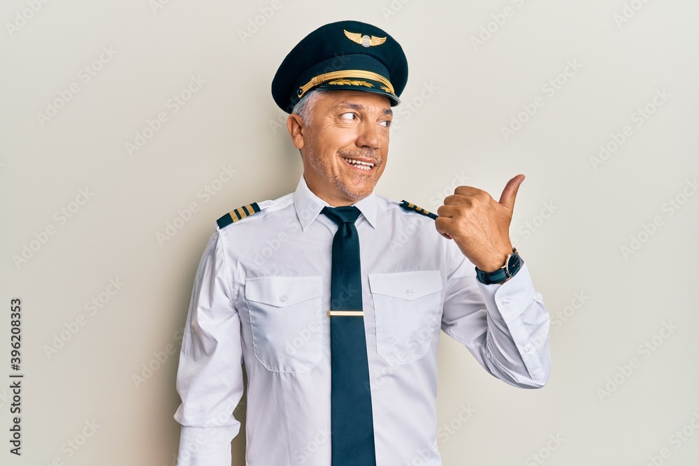 Canvas Prints Handsome middle age mature man wearing airplane pilot uniform smiling with happy face looking and pointing to the side with thumb up.