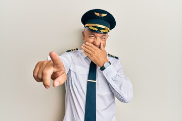 Handsome middle age mature man wearing airplane pilot uniform laughing at you, pointing finger to the camera with hand over mouth, shame expression © Krakenimages.com