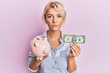 Young blonde girl holding dollar and piggy bank relaxed with serious expression on face. simple and natural looking at the camera.