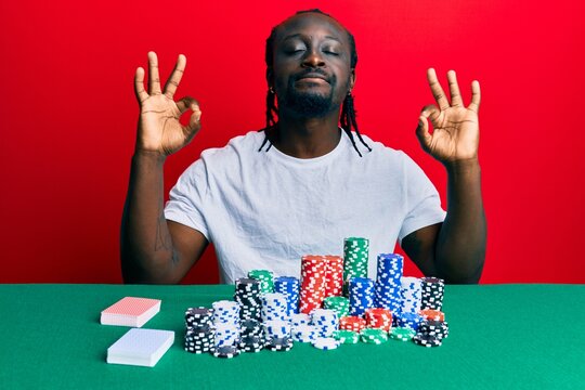 Handsome young black man sitting on the table with poker chips and cards relax and smiling with eyes closed doing meditation gesture with fingers. yoga concept.