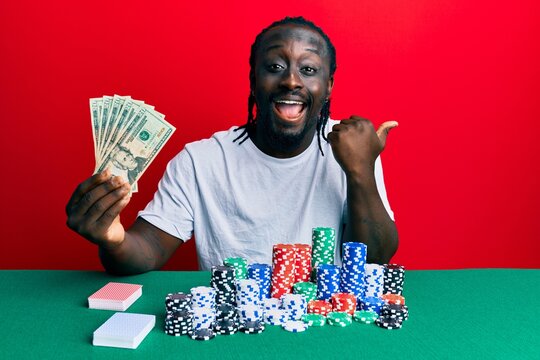 Handsome young black man playing poker holding 20 dollars banknotes pointing thumb up to the side smiling happy with open mouth