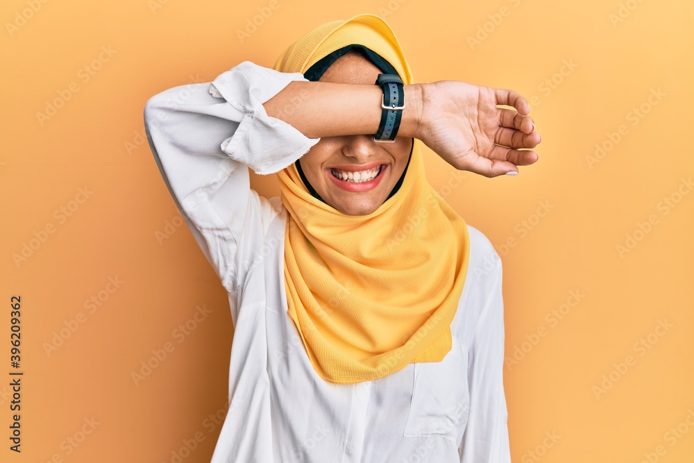 Poster young brunette arab woman wearing traditional islamic hijab scarf covering eyes with arm smiling che