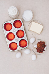 Red paper molds, metal baking tray, eggs, glass of flour, glass of milk, cacao and piece of butter on tablecloth. Ingredients for cupcakes. Top view.