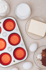 Red paper molds, metal baking tray, eggs, glass of flour, glass of milk, cacao and piece of butter on tablecloth. Ingredients for cupcakes. Top view.