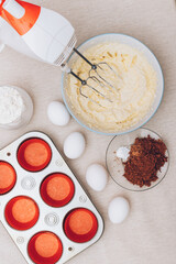Dough for cupcakes in a bowl, red paper molds, metal baking tray, eggs, glass of flour, cacao and mixer on tablecloth. Preparing process to baking.