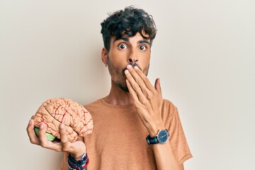 Young hispanic man holding brain covering mouth with hand, shocked and afraid for mistake....