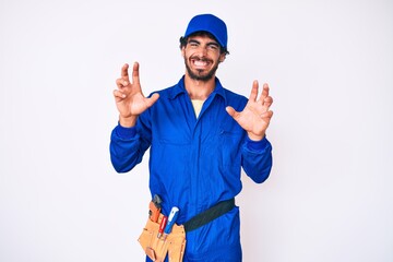 Handsome young man with curly hair and bear weaing handyman uniform smiling funny doing claw gesture as cat, aggressive and sexy expression