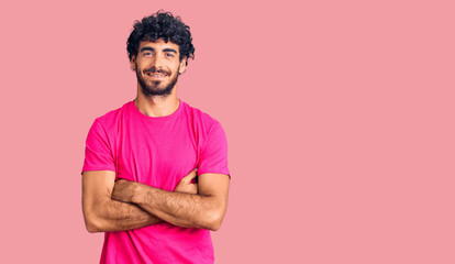 Handsome young man with curly hair and bear wearing casual pink tshirt happy face smiling with crossed arms looking at the camera. positive person.