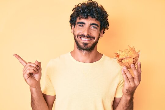 Handsome young man with curly hair and bear holding nachos potato chips smiling happy pointing with hand and finger to the side