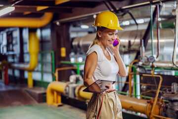 Obraz na płótnie Canvas Middle aged smiling female supervisor in working uniform with helmet on head holding tablet and having phone conversation while standing in heating plant.