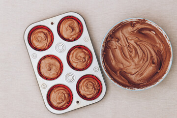 Cacao dough for cupcakes in bowl and red paper molds with dough in metal baking tray on tablecloth. Preparing process to baking. Top view.