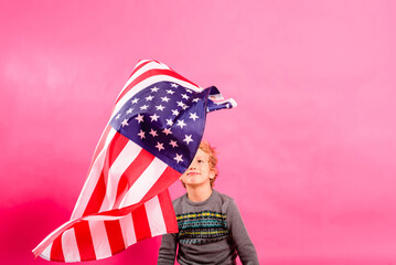 Blond boy under a floating American flag, isolated on a pink background.
