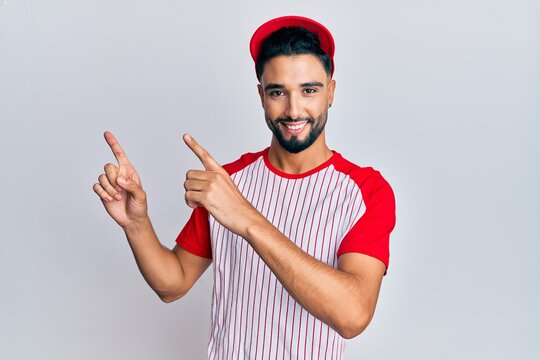 Young man with beard wearing baseball uniform smiling and looking at the camera pointing with two hands and fingers to the side.