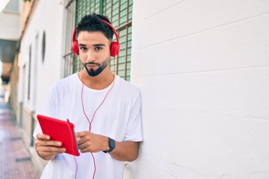 Young arab man with serious expression using headphones and touchpad at the city.