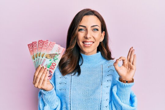 Young brunette woman holding 100 new zealand dollars banknote doing ok sign with fingers, smiling friendly gesturing excellent symbol