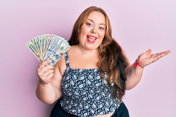 Plus size caucasian young woman holding dollars celebrating achievement with happy smile and winner expression with raised hand