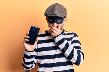 Young handsome bald man wearing burglar mask holding smartphone covering mouth with hand, shocked and afraid for mistake. surprised expression