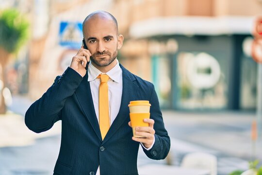Young hispanic bald businessman with serious expression talking on the smartphone drinking coffee at the city